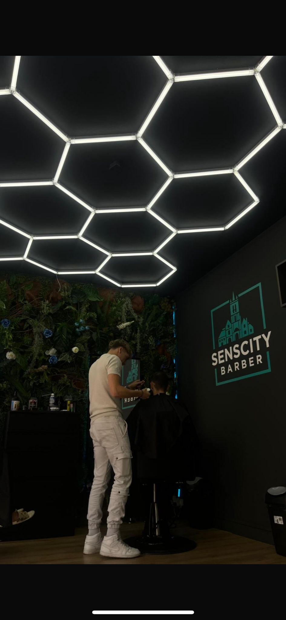 Un barbier au travail sous les néons hexagonaux du salon, devant le mur au logo Sens City Barber